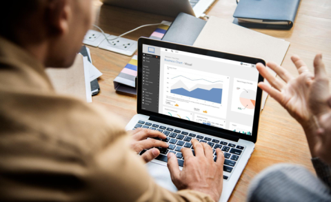 Two people working together; one is typing on the laptop with charts on the screen while the other is giving out gestures.